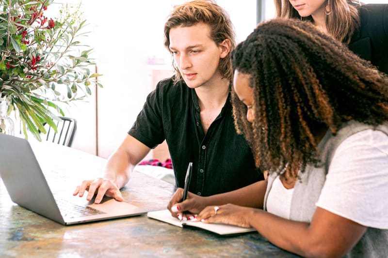 Two students collaborating at a table, one typing on a laptop and the other taking notes