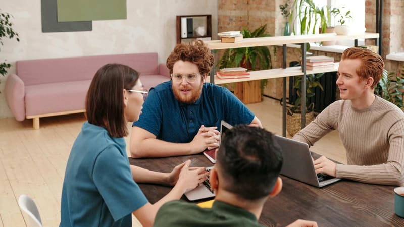 Four students talking around a table with laptops in a bright, plant-filled room